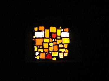 Quadratisches Buntglasfenster, gelb und orange, in Dunkelheit.